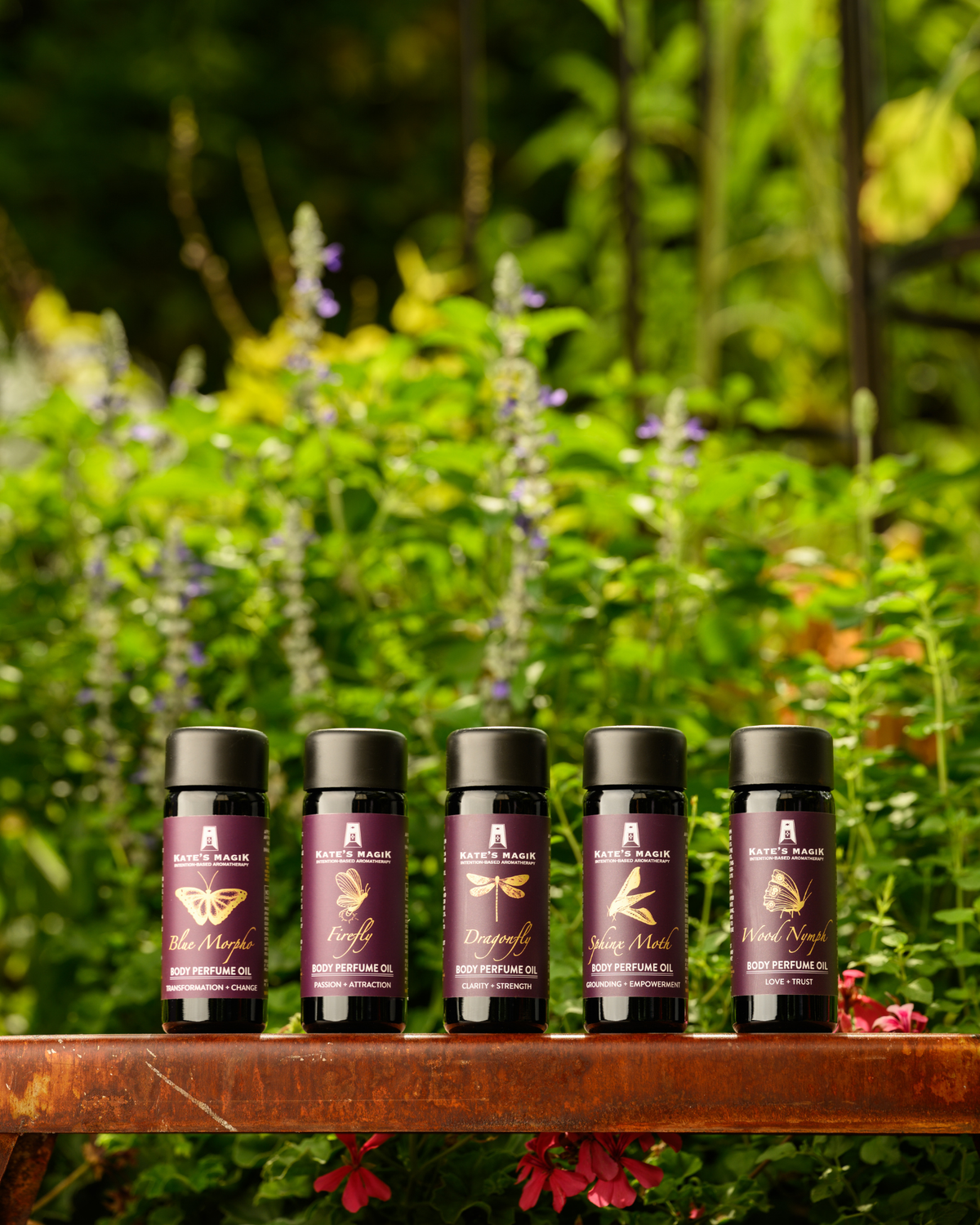 Five 100ml violet glass bottles of Kate's Magik Body Perfume OIl with purple labels, gold and white lettering and black caps, lined up on a rust colored metal stand in a lush green garden.