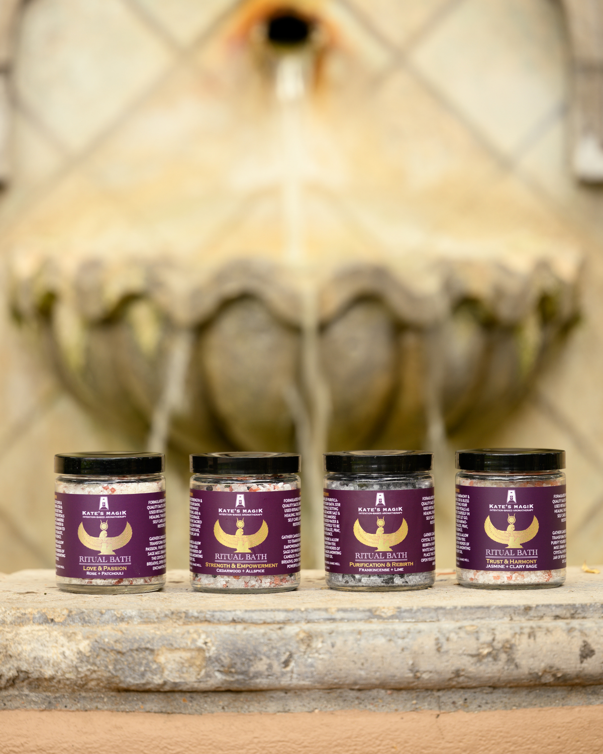 Four jars of Ritual Bath stands arranged in a row on a stone ledge with a blurred fountain in the background.
