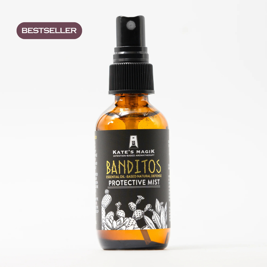 An amber glass spray bottle labeled 'Kate's Magik Banditos Protective Mist' is centered on a white background, surrounded by botanical props including a sliced lemon, sage leaves, rosemary, cinnamon sticks, and clove. No people or real environment are present, so this is Product/Packaging rather than Lifestyle.