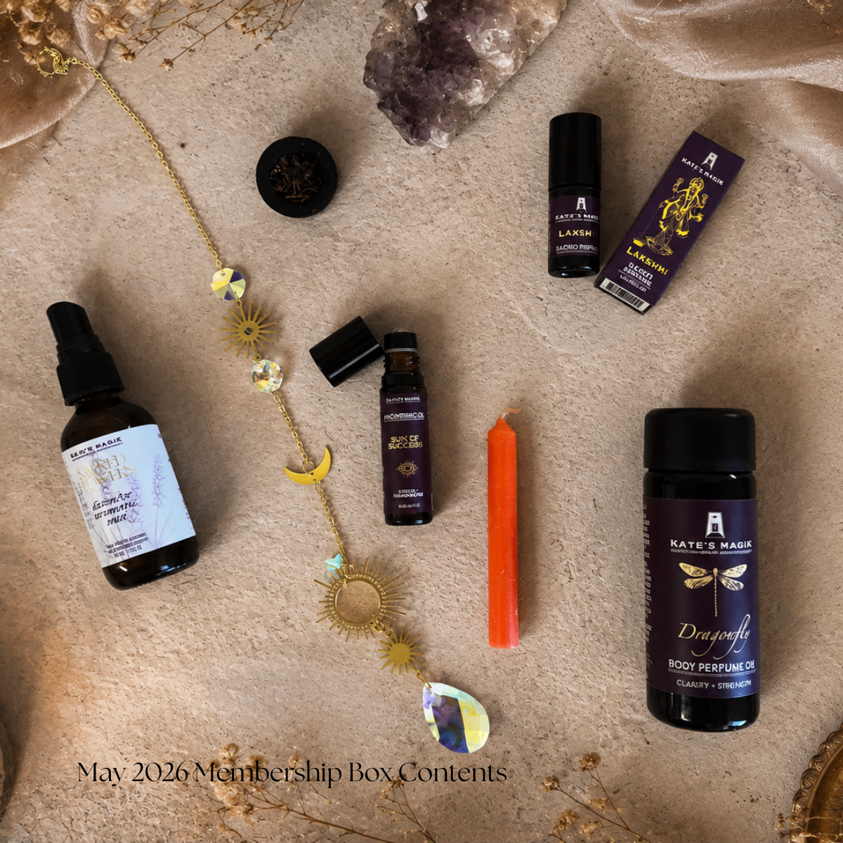 A styled flat-lay of Kate Magik oils and body care products on a beige fabric background with crystals and dried flowers. Visible items include a lavender body mist bottle, a Sun of Success Anointing Oil roll-on bottle, a dragonfly body perfume oil bottle, a small amber candle, a gold-toned sun catcher with moon and sun charms, and an incense disk.