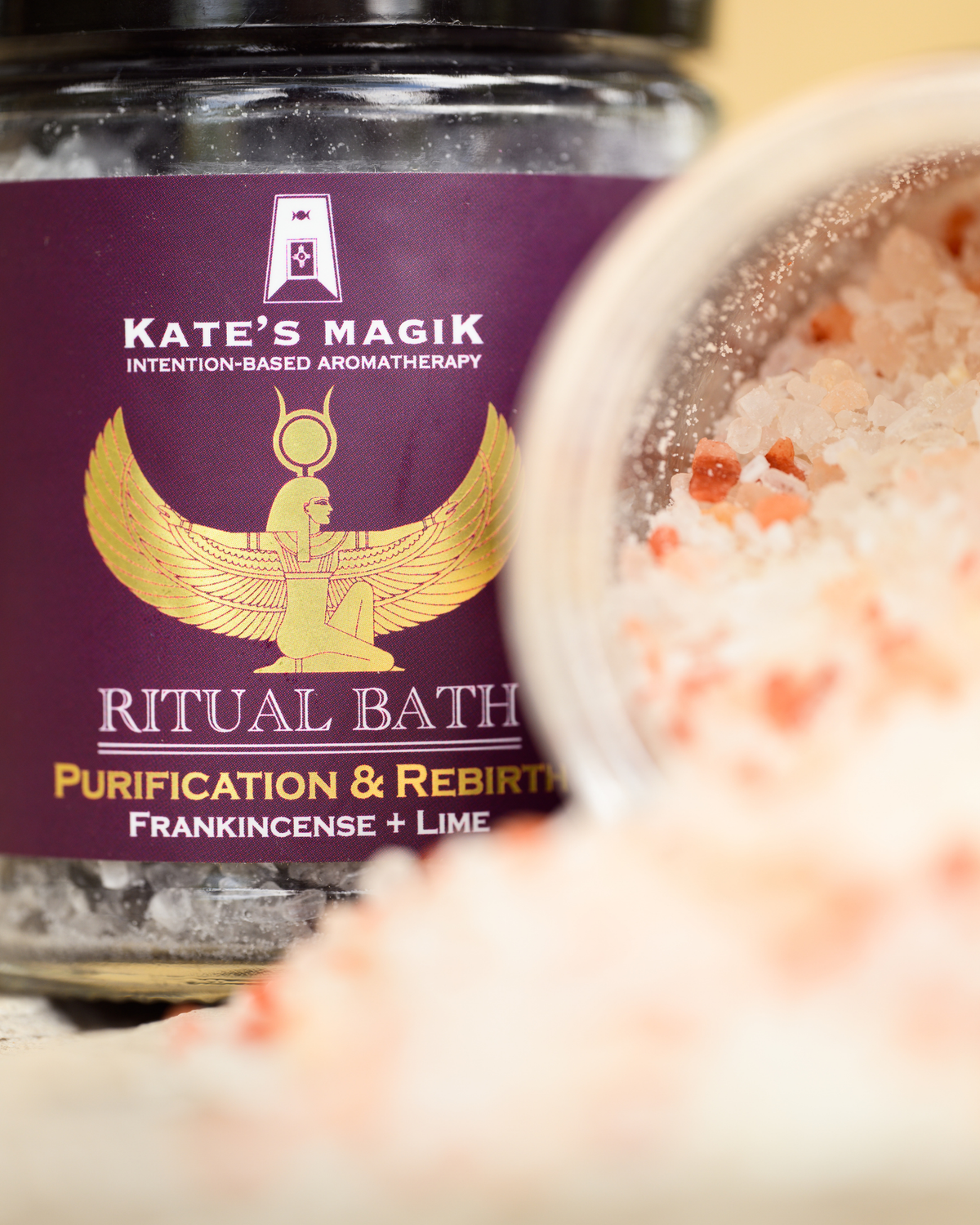 A close-up of a glass jar labeled 'Kate's Magik Ritual Bath Purification & Rebirth Frankincense + Lime' with a deep purple label and gold emblem, alongside spilled pink bath salts in the foreground.