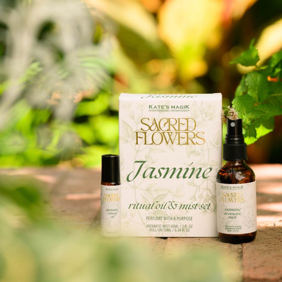 Kate's Magik Sacred Flowers Jasmine ritual oil and mist set, featuring a white box with gold and green colored lettering, a small roller ball bottle, and a 2oz amber spray bottle, styled outdoors on a stone surface with a blurred green garden background.