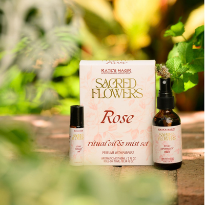 Kate's Magik Sacred Flowers Rose ritual oil and mist set, featuring a white box with gold and rose colored lettering, a small roller ball bottle, and a 2oz amber spray bottle, styled outdoors on a stone surface with a blurred green garden background.