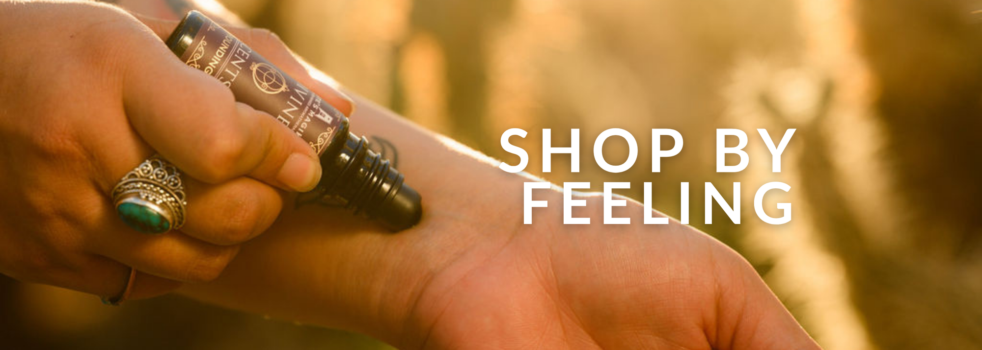 A close-up of a hand holding a small cylindrical roll-on bottle over another hand, with large white text reading 'SHOP BY FEELING' on the right. The background is a warm, blurred outdoor scene in golden tones.