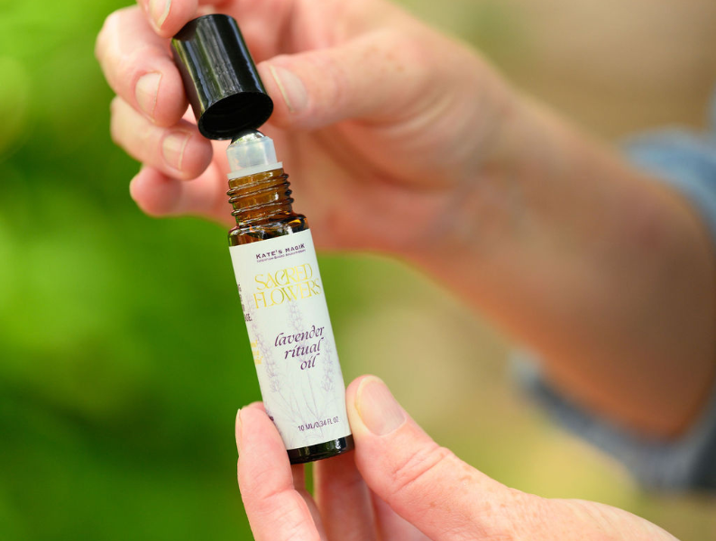 A person holds an amber glass bottle of Kate's Magik Lavender Ritual Oil, unscrewing the black cap, against a soft, blurred green and beige background.