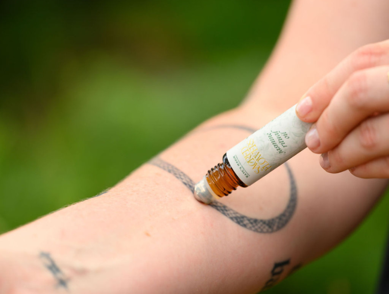 A close-up of a hand holding a small amber glass roller bottle over a person's forearm with a circular tattoo, against a blurred green outdoor background.