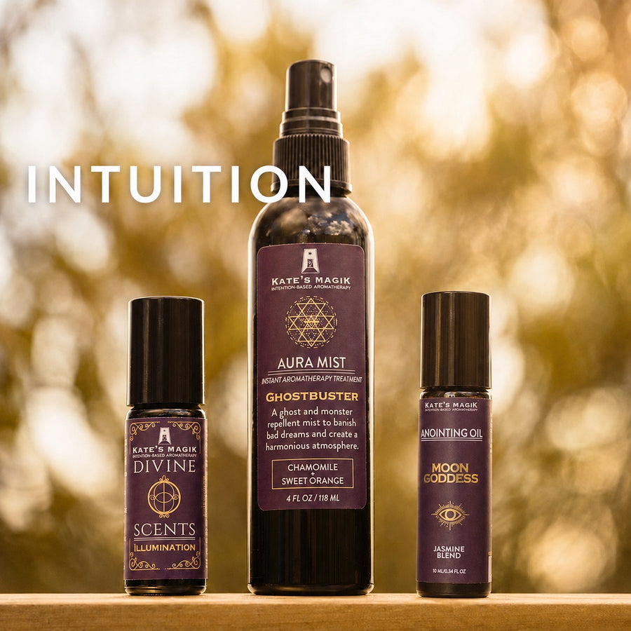 Three Kate's Magick essential oil and aromatherapy products displayed on a wooden surface against a warm, blurred outdoor background. The products include a central Aura Mist spray bottle, a Divine Scents roll-on bottle, and a Noon Goddess roll-on bottle, with the word 'INTUITION' written in white text on the left.